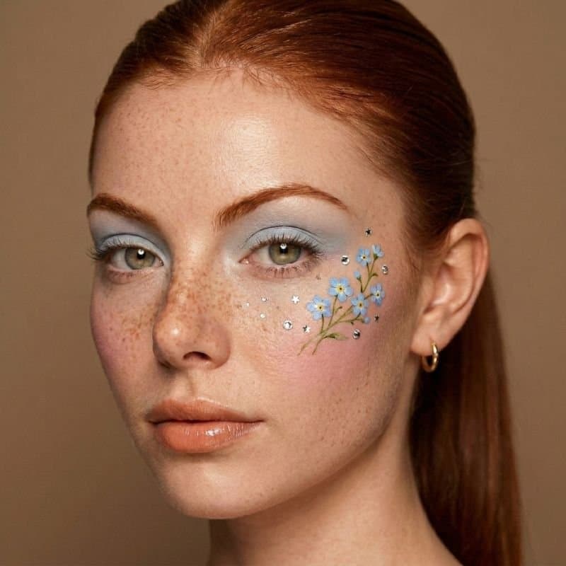 Nahaufnahme einer jungen Frau mit roten Haaren und Sommersprossen. Sie trägt blaues Augen-Make-up und kleine blaue Blumen sowie silberne Sterne auf der Wange: Beispiel für feine Hautstruktur in KI-Bildern.